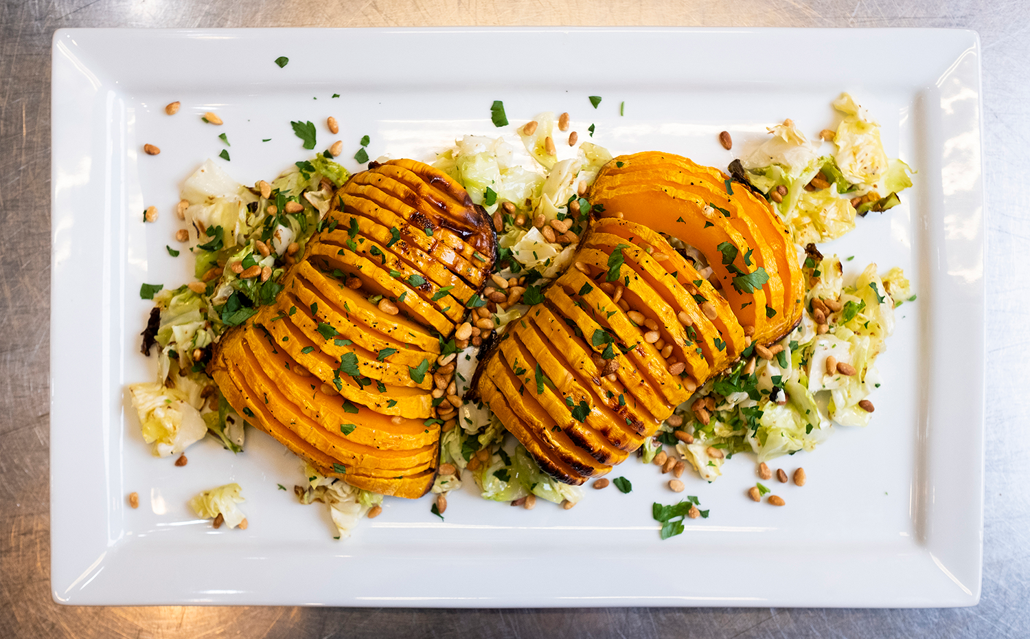 Overhead view of two hasselback-sliced roasted butternut squash halves served over a bed of chopped roasted cabbage, garnished with toasted pine nuts and fresh parsley on a white rectangular platter.