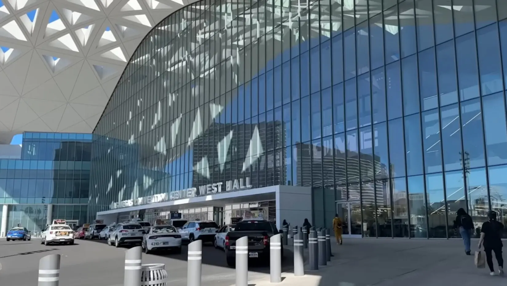 A bright, sunny daytime view of the modern glass exterior of the Las Vegas Convention Center West Hall, with cars dropping off attendees near the entrance for the International Pizza Expo.