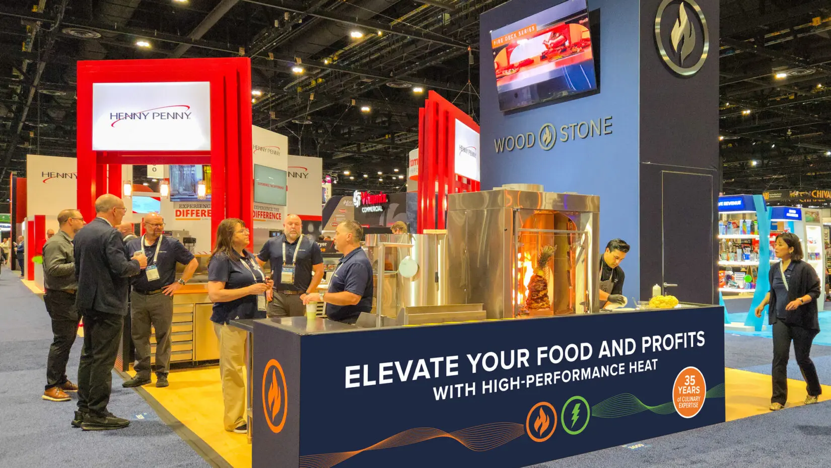 A bustling trade show booth for Wood Stone and Henny Penny at the National Restaurant Association Show, featuring a towering blue display with the Wood Stone flame logo, an active Fire Deck series stone hearth oven, and staff engaging with attendees.