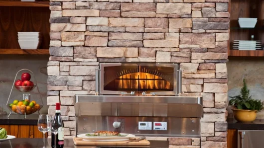A straight-on view of a built-in Wood Stone Bistro Home 4343 oven seamlessly integrated into a floor-to-ceiling stacked stone pillar in an indoor kitchen, featuring a stainless steel face, glass doors revealing a live fire, and a curved dark granite dining counter in the foreground.
