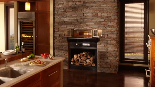 A wide view of a warm, luxurious indoor kitchen featuring a built-in Wood Stone home oven seamlessly integrated into a floor-to-ceiling dark stacked stone wall, complete with open glass doors revealing a live fire, a black lower panel with firewood storage, and an adjacent kitchen island prepped with a fresh pizza.