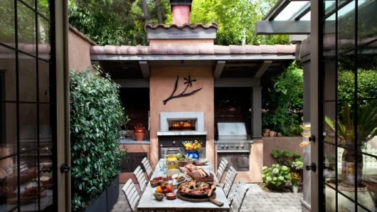 A view looking through open glass French doors into a lush outdoor courtyard featuring a long dining table laden with food, backed by a built-in Wood Stone Bistro Home 4343 oven enclosed in a custom terracotta stucco facade with a tiled roof, adjacent to a built-in stainless steel grill.