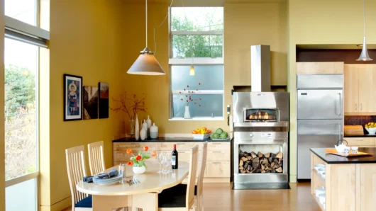 A wide view of a bright, modern indoor residential kitchen featuring a freestanding, stainless steel Wood Stone Bistro Home 4343 oven with a prominent exhaust hood and a lower firewood storage cavity, set against warm yellow walls adjacent to a dining table and light wood cabinetry.