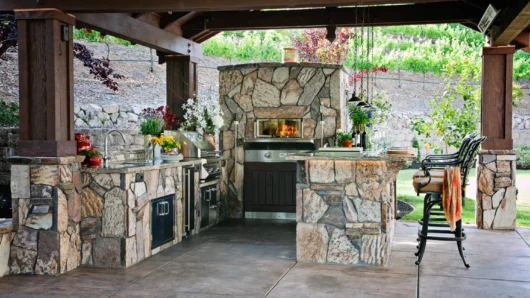 A wide view of a luxurious covered outdoor kitchen pavilion featuring a built-in Wood Stone home oven seamlessly enclosed in a massive rustic fieldstone facade, matching stone pillars, and countertops, complete with a built-in grill, sink, and bar seating overlooking a terraced garden.