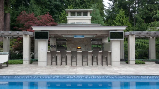 A wide architectural view of a luxurious outdoor poolside kitchen pavilion featuring a built-in Wood Stone home oven clad in stone, centrally located behind a long counter with six bar stools, and flanked by mounted flat-screen televisions.