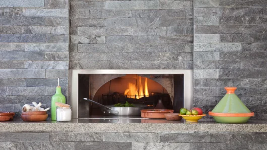 A straight-on view of a built-in Wood Stone home oven seamlessly integrated into a modern dark grey stacked stone wall, featuring a live fire cooking food in a skillet, with a granite mantle displaying a vibrant green tagine, fresh garlic, and olive oil.