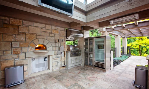A wide view of a luxurious covered outdoor kitchen featuring a Wood Stone home oven built into a light tan stone wall with an arched opening, flanked by hanging pizza peels, a built-in stainless steel grill, and a large glass-door refrigerator beneath a wooden beam ceiling.
