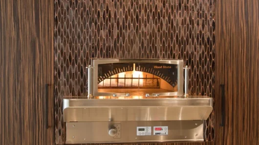 A straight-on view of a built-in Wood Stone Bistro Home oven seamlessly integrated into sleek, dark wood-grain cabinetry. The stainless steel oven features a glass viewing door revealing a live fire, surrounded by a textured, copper-toned diamond mosaic tile backsplash.