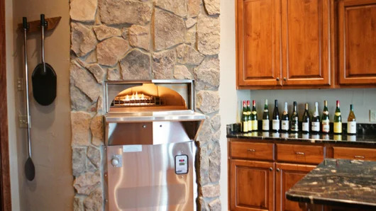 A view of an elegant indoor home kitchen featuring a Wood Stone oven built into an angled corner wall covered in a floor-to-ceiling rustic stone facade, contrasting with the oven's stainless steel front, adjacent warm wood cabinetry, and dark granite countertops.