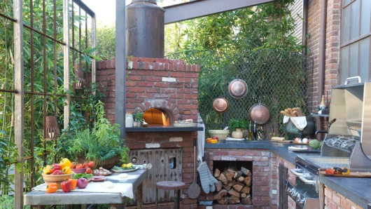 A sprawling outdoor kitchen patio featuring a built-in Wood Stone Mountain Series oven fully enclosed in rustic red brick, complete with an arched opening, a tall metal exhaust chimney, adjacent built-in firewood storage, and a stainless steel prep table loaded with fresh vegetables.