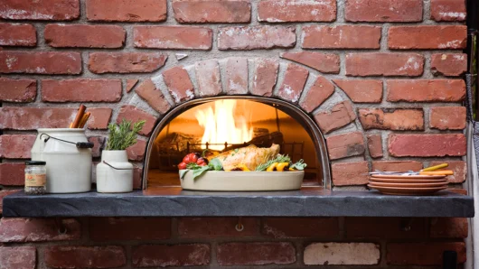 A close-up view of a built-in Wood Stone home oven featuring a custom traditional red brick facade and an arched opening. A live fire glows inside, while a freshly roasted chicken and vegetables sit on a dark stone mantle in the foreground alongside rustic cooking utensils, fresh herbs, and stacked plates.
