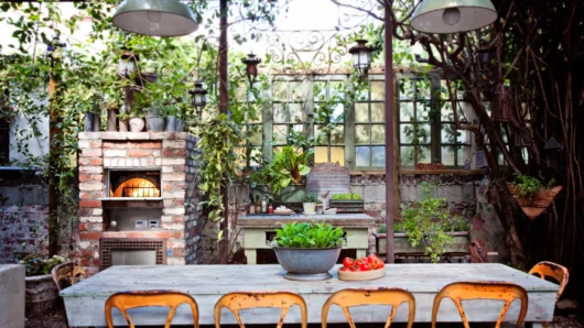 A rustic outdoor garden patio featuring a built-in Wood Stone Bistro Home oven enclosed in weathered red and white brick, surrounded by lush potted plants, vintage paned windows, and a weathered wooden dining table with distressed orange metal chairs.