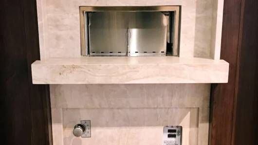 A straight-on view of a built-in Wood Stone oven featuring a luxurious light cream marble facade and built-in mantle, contrasted against dark wood paneling, with a stainless steel night door covering the cooking opening and seamlessly integrated lower controls.