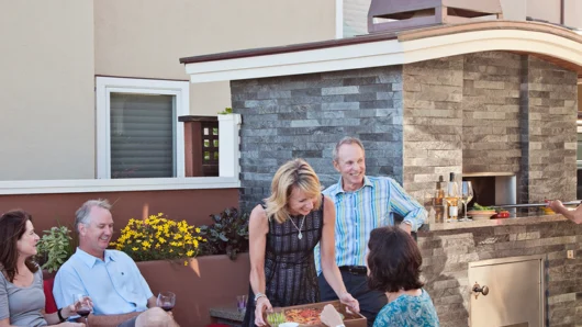 A lively outdoor patio scene featuring a group of adults socializing and being served freshly baked pizza next to a built-in Wood Stone home oven, which is fully enclosed in a dark grey stacked stone facade with a stainless steel lower access panel and a large metal exhaust chimney.